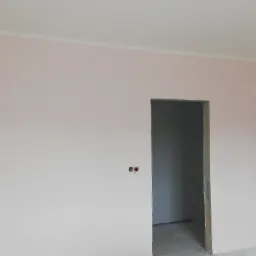 Świeżo pomalowane, jasnoróżowe ściany w pokoju z widocznymi otworami po gniazdkach elektrycznych i surowym otworem drzwiowym.