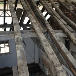 Drewniana konstrukcja dachu w trakcie remontu, z widocznymi belkami, oknem w tle i fragmentami ceglanego komina.