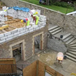 Budowa domu w trakcie: murowane ściany z bloczków i kamienia, drewniany szkielet dachu, robotnicy w żółtych kamizelkach, betonowe schody zewnętrzne, betoniarka, widok z góry.