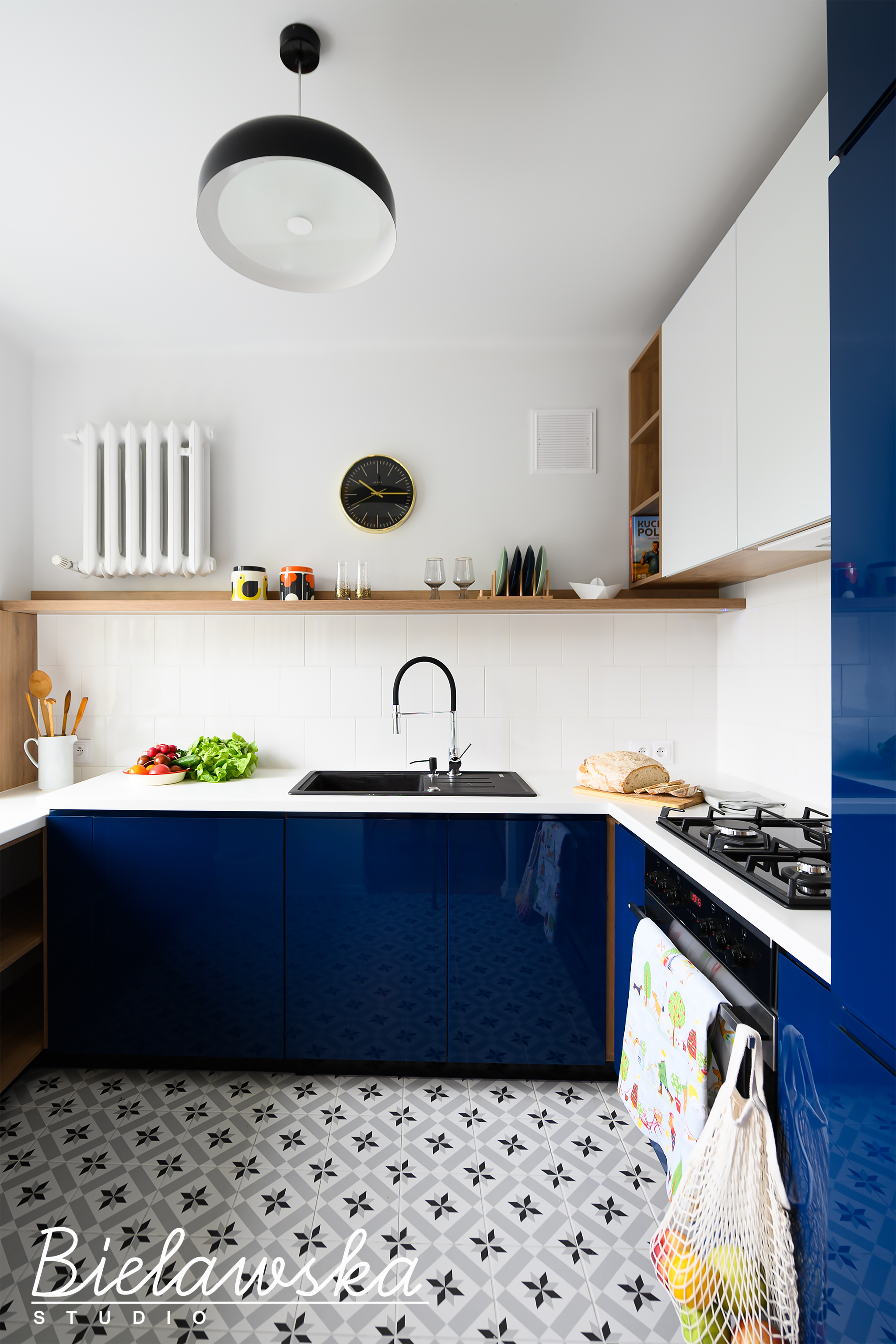 Nowoczesna kuchnia z granatowymi szafkami na tle wzorzystej podłogi, białe płytki nad blatem, drewniane półki z dekoracjami i świeżymi warzywami, czarny zlew i bateria, wisząca lampa w stylu retro.