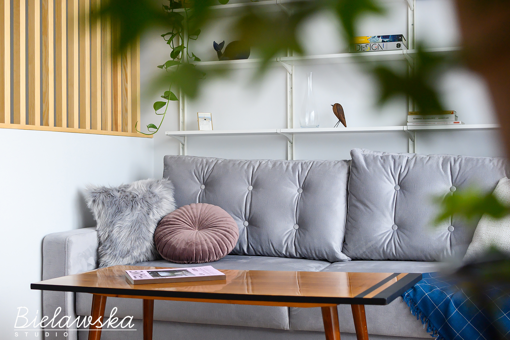 Szary narożnik z poduszkami: futrzaną i okrągłą, stolik kawowy z książką, białe półki z dekoracjami i drewniane lamele na ścianie, widok przez roślinę doniczkową.