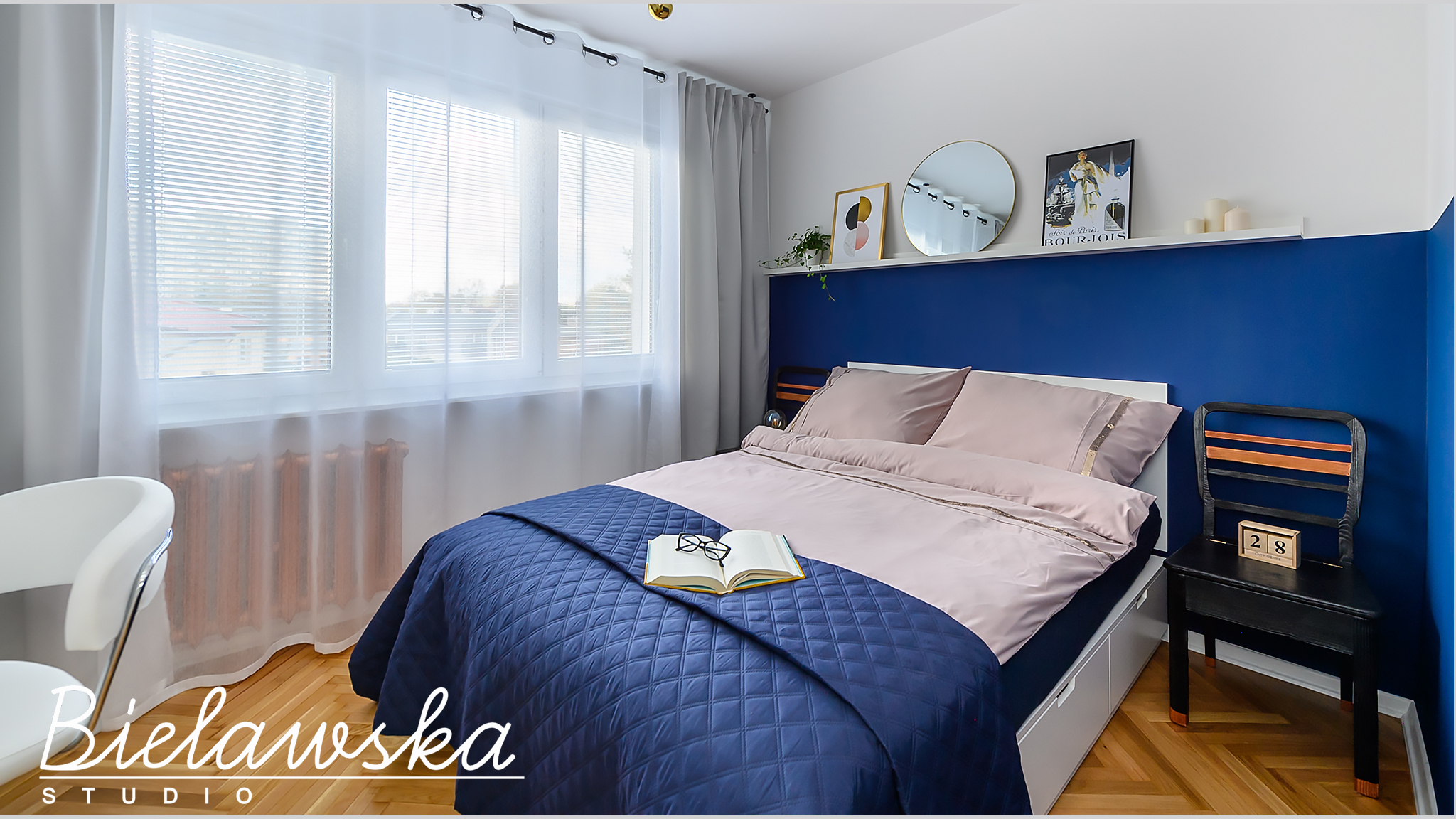 Nowoczesna sypialnia z łóżkiem przykrytym granatową kołdrą, otwartą książką i okularami, niebieska ściana za łóżkiem, drewniana podłoga w jodełkę.