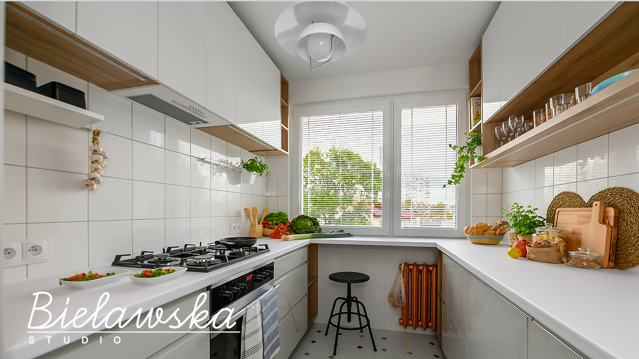 Jasna kuchnia z białymi płytkami, drewnianymi półkami, czarną kuchenką gazową i dodatkami spożywczymi, widok na okno z zielenią za szybą, z logo firmy w lewym dolnym rogu.