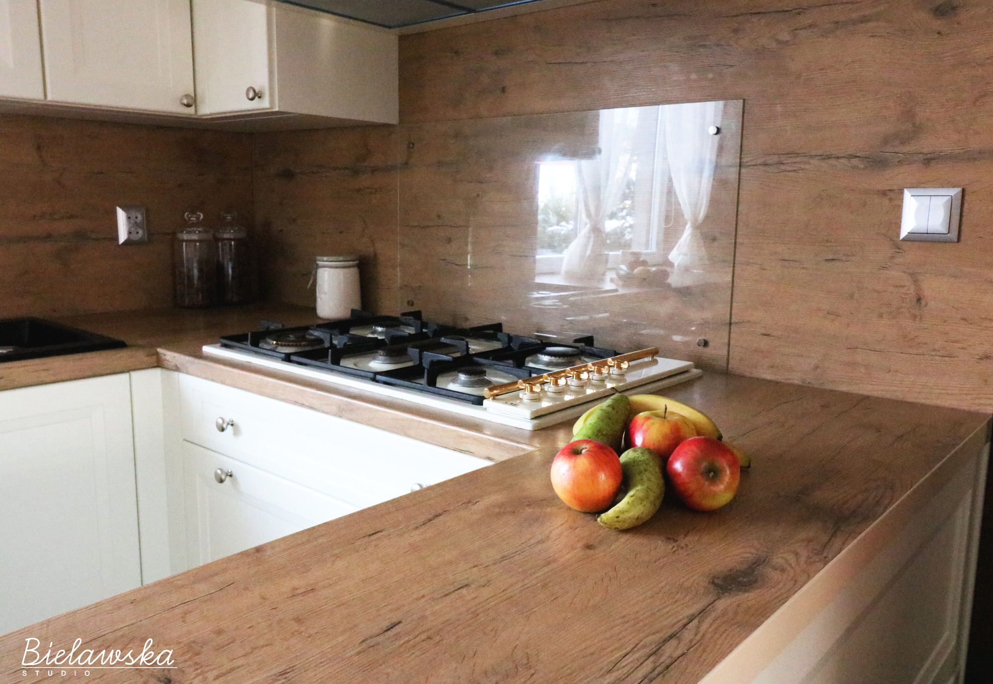 Klasyczna kuchnia z drewnianym blatem, białymi szafkami i owocami na blacie, widok na okno z firankami w tle.
