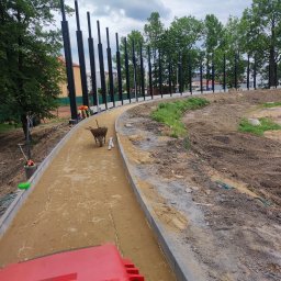 ADAMIEC - Budowa ścieżki parkowej: wyrównany piasek, obrzeża z kostki, w tle czarne słupy konstrukcji i zieleń. Widoczna taczka i zagęszczarka. Prace w toku.