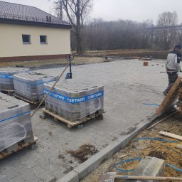 ADAMIEC - Ułożona szara kostka brukowa z paletami kostki Betard w tle, widoczny fragment budynku i pracownik w trakcie pracy. Krawężnik oddziela kostkę od ziemi.