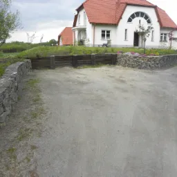 Ukształtowanie terenu wokół domu z wykorzystaniem kamiennych i drewnianych murków oporowych, widoczny podjazd z szutru i trawnik przed budynkiem.