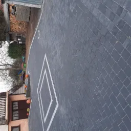 Ułożona szara kostka brukowa z białymi liniami wyznaczającymi pole gry, widok z góry, w tle budynek mieszkalny i składowane drewno.