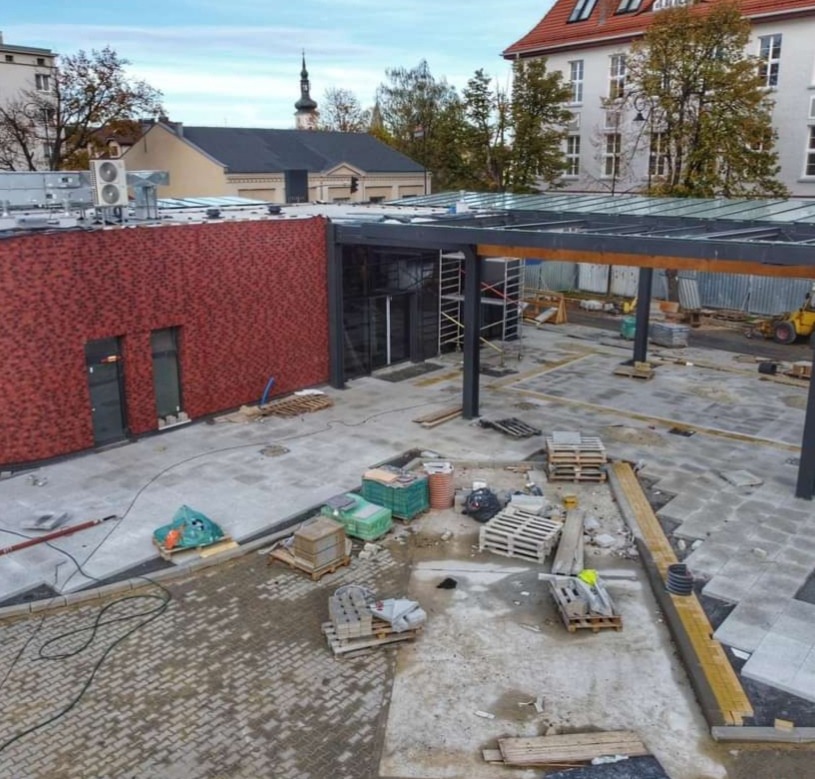 Plac z częściowo ułożoną kostką brukową, widoczne palety z kostką, budynek z czerwoną elewacją i zadaszenie. Prace brukarskie w toku, widok z góry.