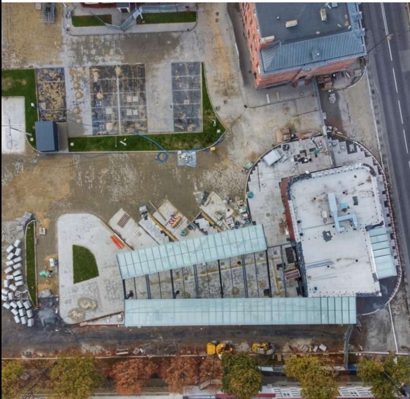 Widok z góry na plac budowy z elementami zagospodarowania terenu: przygotowane podłoże, fragmenty zieleni, budynki w tle i maszyny budowlane. Ujęcie z drona.