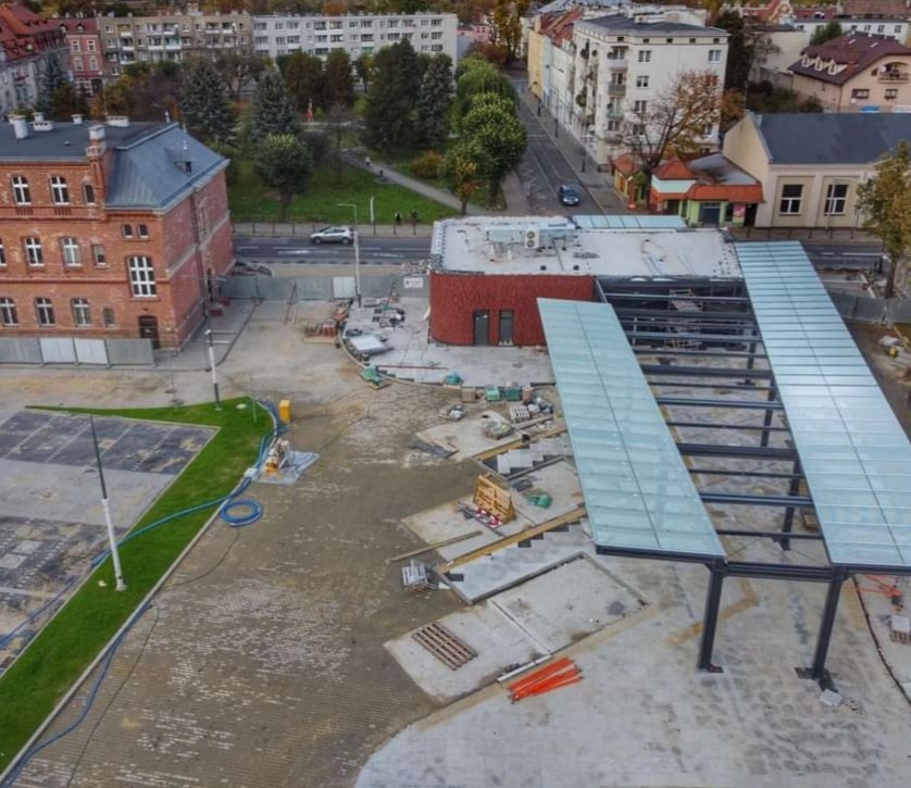 Plac budowy z widoczną konstrukcją zadaszenia, świeżo ułożoną kostką i fragmentem trawnika. W tle budynki miejskie i drzewa. Ujęcie z drona.