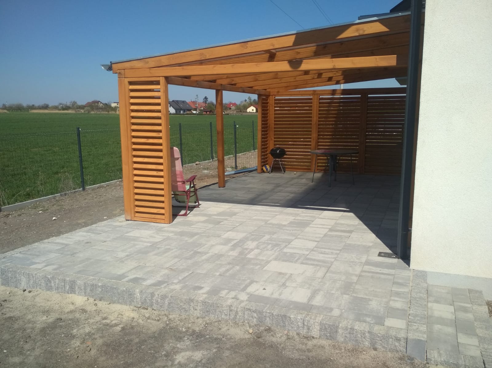 Zadaszona, drewniana altana z ażurowymi ścianami, wyposażona w grill i stolik, stoi na wyłożonym kostką brukową tarasie. W tle widoczne zielone pole i zabudowania.