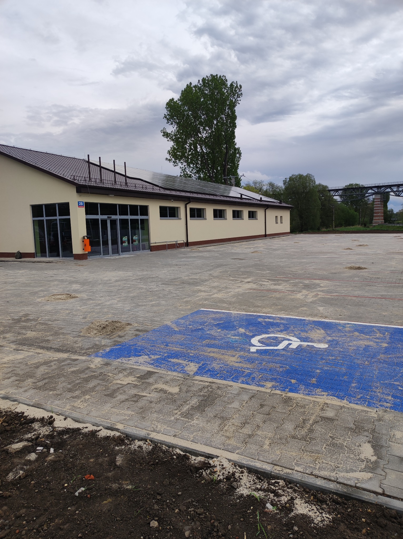 Nowy parking z kostki brukowej z wyznaczonym miejscem dla osób niepełnosprawnych, budynek w tle, pochmurne niebo. Widoczny fragment mostu w oddali.