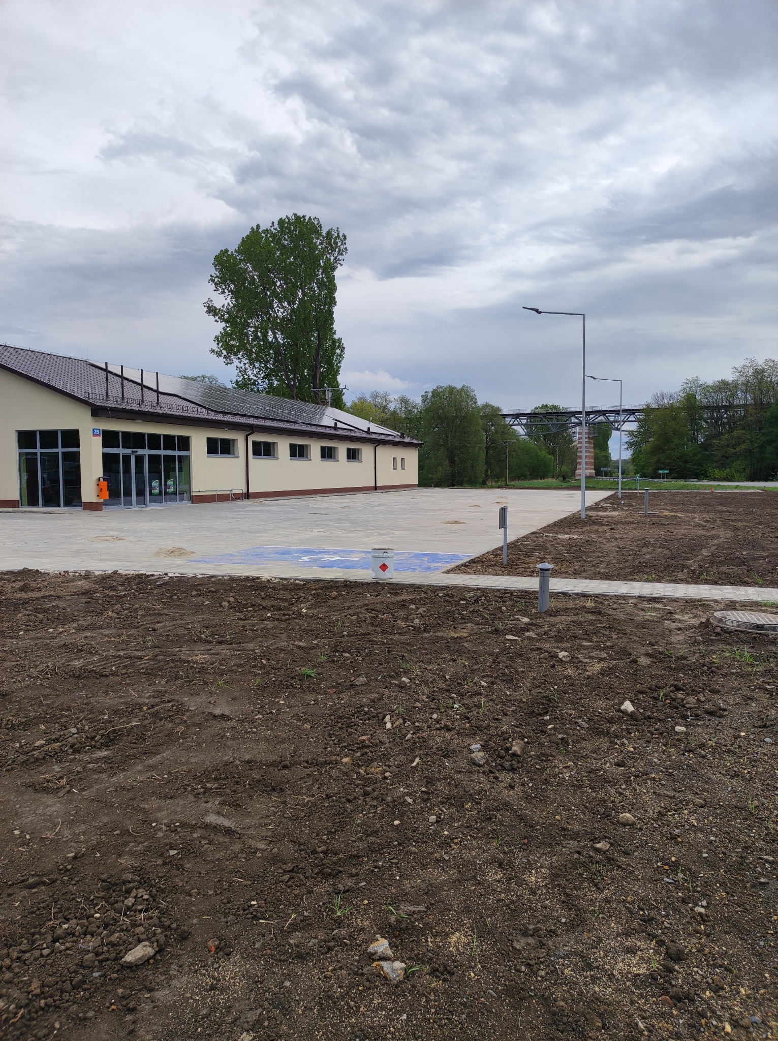 Nowo wybudowany budynek z parkingiem dla niepełnosprawnych i przygotowanym terenem pod zieleń, widok na wiadukt w tle.