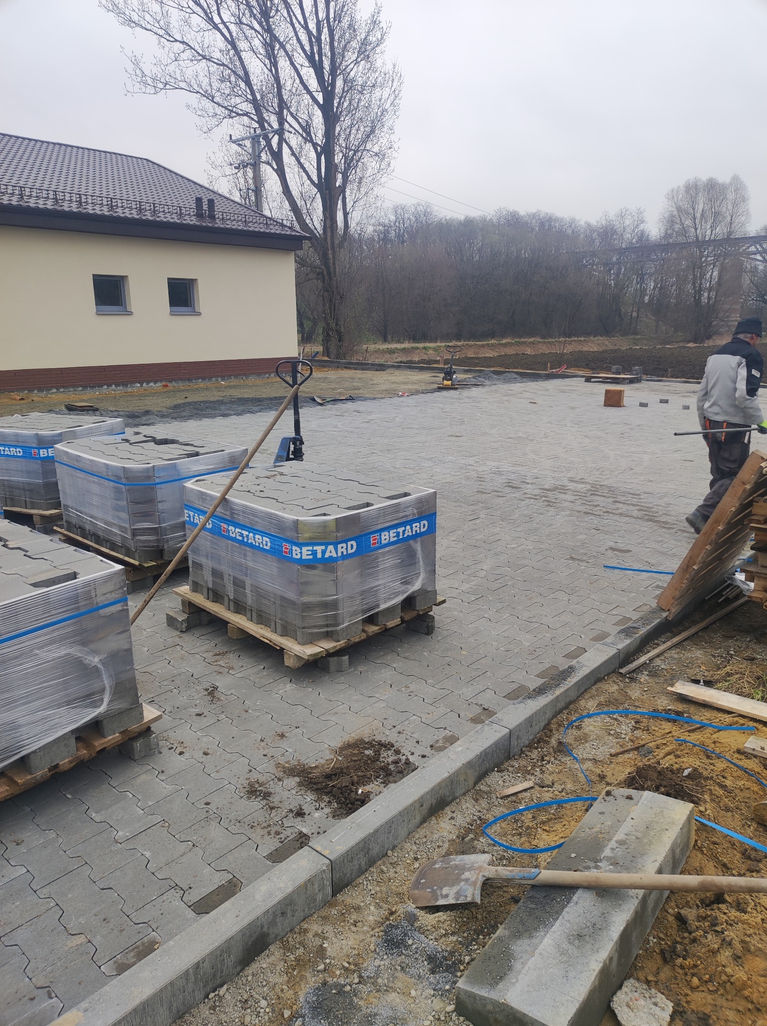 Ułożona szara kostka brukowa z paletami kostki Betard w tle, widoczny fragment budynku i pracownik w trakcie pracy. Krawężnik oddziela kostkę od ziemi.