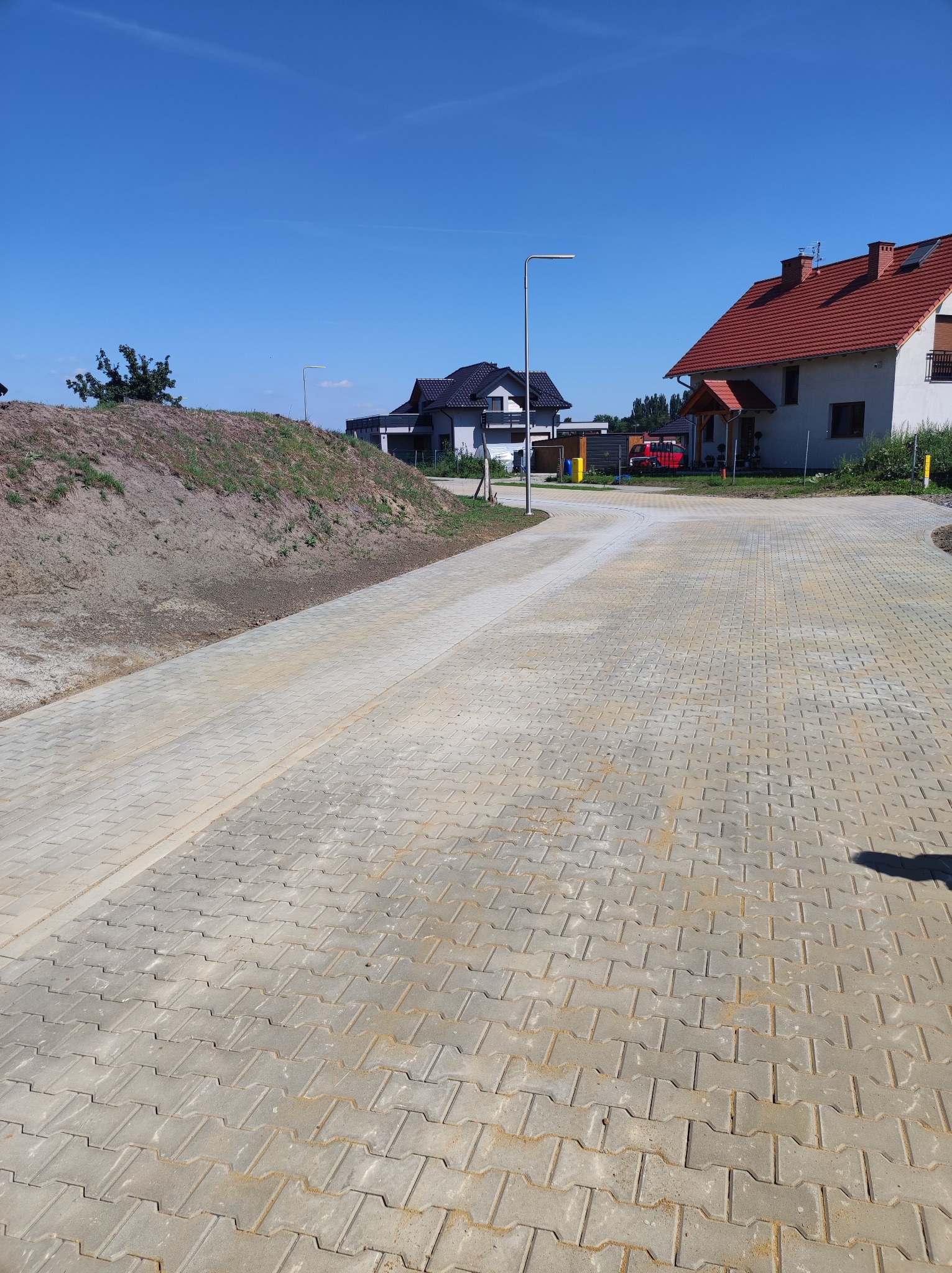 Jasnoszara kostka brukowa ułożona na podjeździe do domu z czerwoną dachówką, w tle widać świeżo usypaną ziemię i inne budynki. Słoneczny dzień.