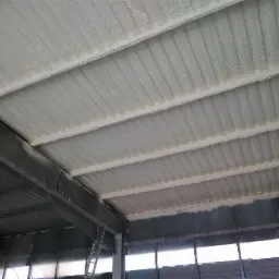 Metalowa konstrukcja dachu hali przemysłowej pokryta warstwą izolacyjną z pianki, widoczny fragment ściany z oknami i drabina oparta o filar.