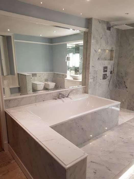 Nowoczesna łazienka wykończona białym marmurem, z zabudowaną wanną, lustrem nad wanną odbijającym toaletę i bidet, oraz prysznicem w kabinie walk-in.