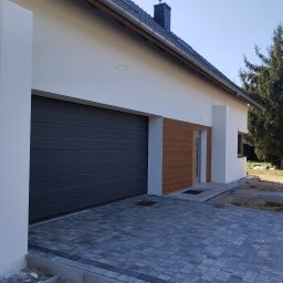 MiKO COMPLEX MICHAŁ KOZŁOWSKI - Nowoczesny dom z grafitową bramą garażową, elementami drewnianej elewacji i wybrukowanym podjazdem z kostki w odcieniach szarości.