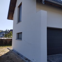 MiKO COMPLEX MICHAŁ KOZŁOWSKI - Świeżo otynkowana, jasna elewacja domu jednorodzinnego z ciemnymi oknami i grafitową bramą garażową, widok z boku na tle zieleni.