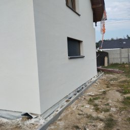 MiKO COMPLEX MICHAŁ KOZŁOWSKI - Świeżo otynkowana, biała elewacja domu jednorodzinnego z widoczną siatką drenażową przy fundamentach i częściowo odsłoniętą izolacją przeciwwilgociową, okno z zewnętrznymi roletami.