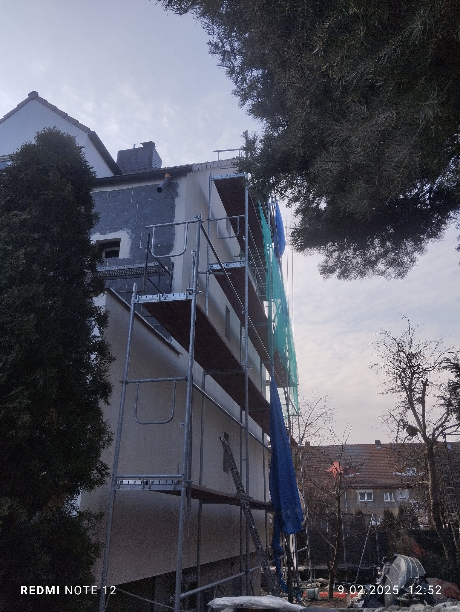 Budynek w Miłosławiu w trakcie prac elewacyjnych, widoczne rusztowanie, fragment ocieplonej ściany i fragment ściany po nałożeniu tynku.