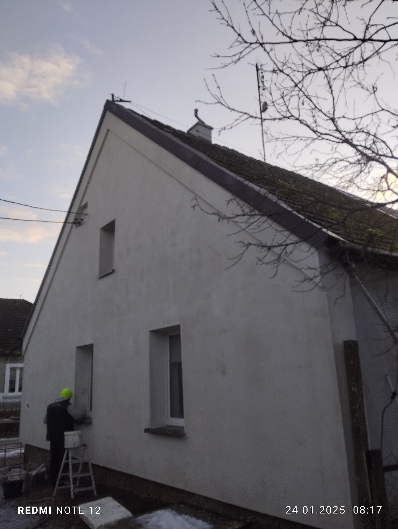 Tynkowanie elewacji domu jednorodzinnego z użyciem drabiny, widoczny pracownik w żółtym kasku i wiadro z zaprawą, elewacja w trakcie prac wykończeniowych, pochmurny dzień.