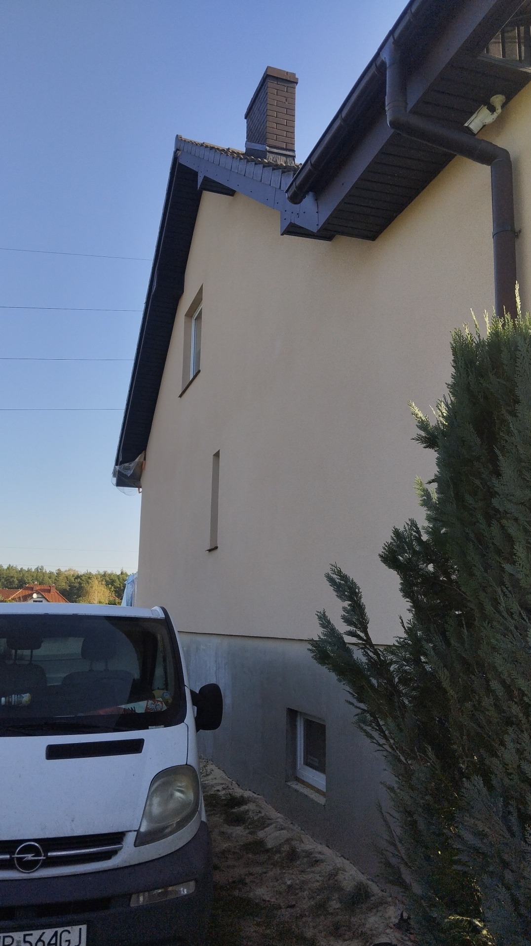 Fragment elewacji domu po nałożeniu jasnego tynku, widoczny samochód dostawczy i fragment iglaka