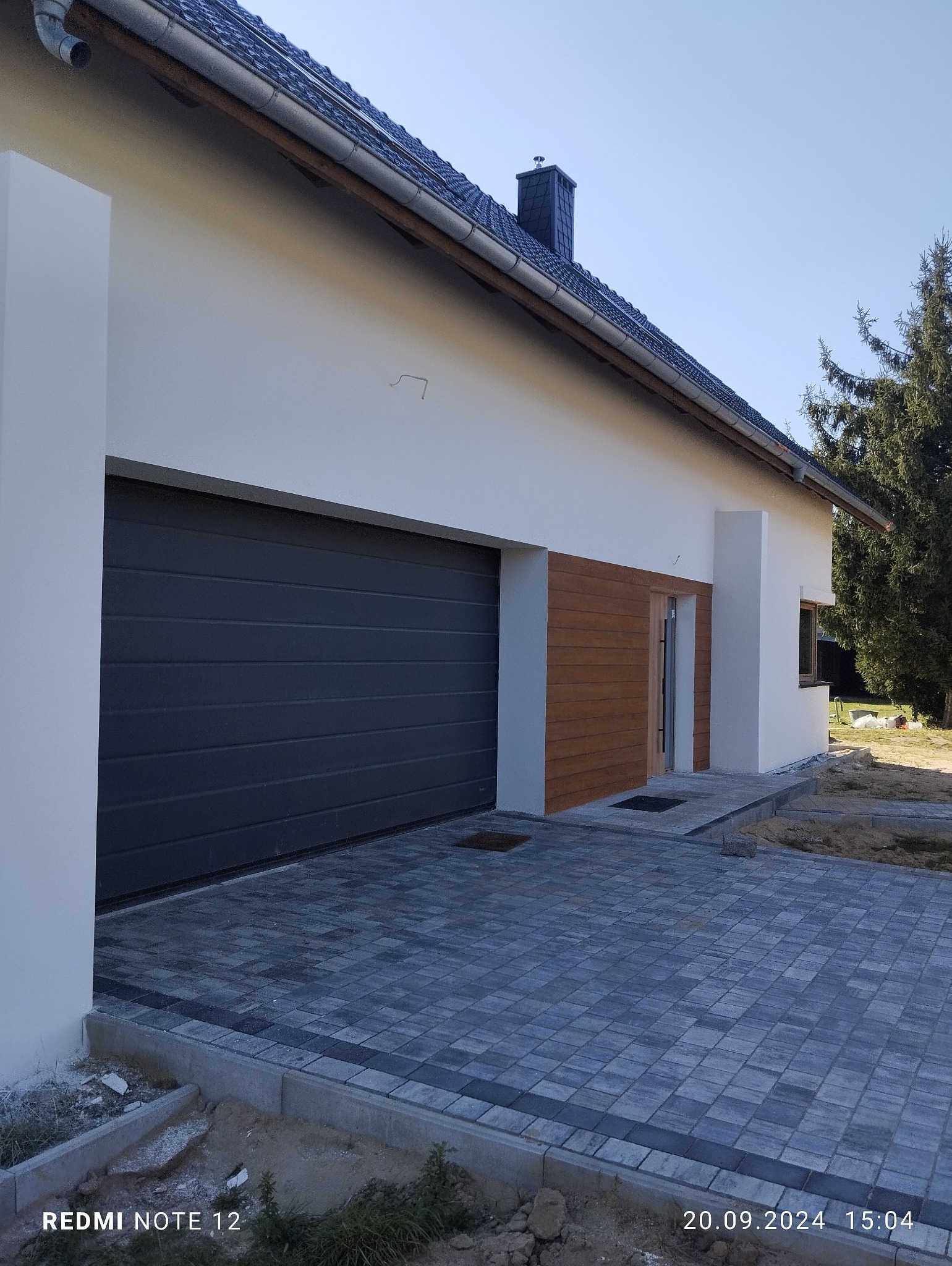 Nowoczesny dom z grafitową bramą garażową, elementami drewnianej elewacji i wybrukowanym podjazdem z kostki w odcieniach szarości.