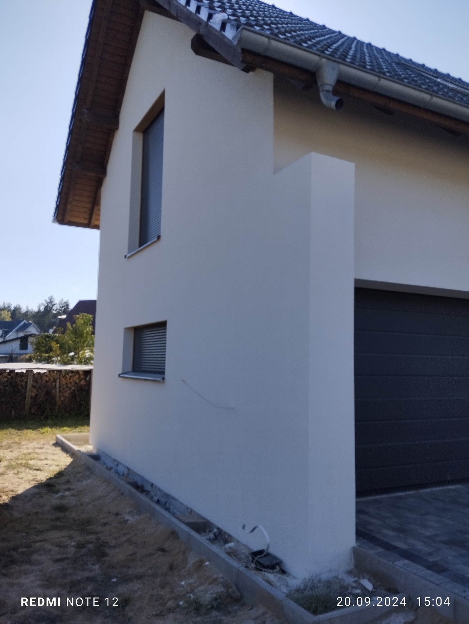 Świeżo otynkowana, jasna elewacja domu jednorodzinnego z ciemnymi oknami i grafitową bramą garażową, widok z boku na tle zieleni.