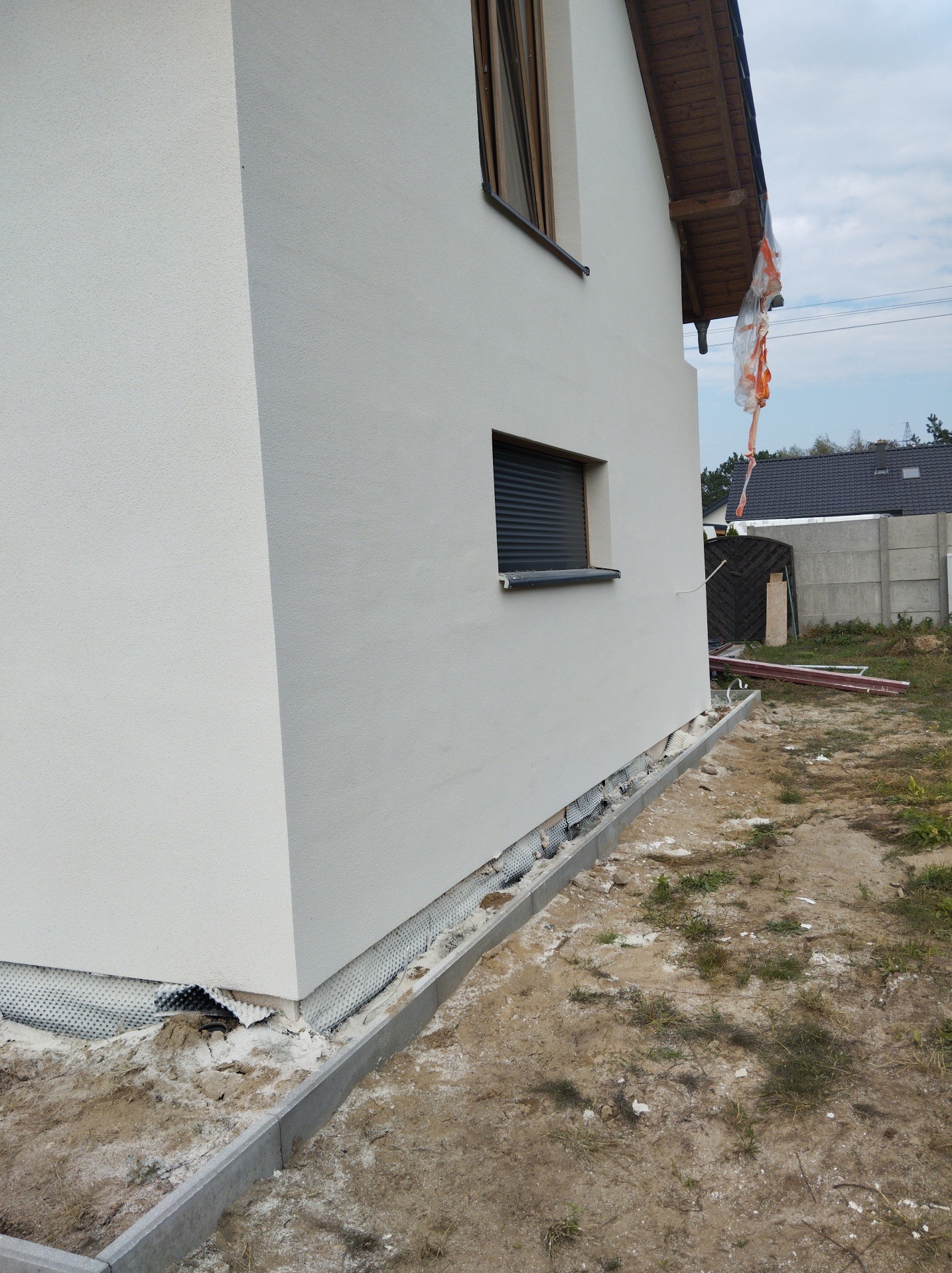 Świeżo otynkowana, biała elewacja domu jednorodzinnego z widoczną siatką drenażową przy fundamentach i częściowo odsłoniętą izolacją przeciwwilgociową, okno z zewnętrznymi roletami.