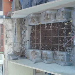 Przygotowanie do wylewki fundamentów: zbrojenie siatką stalową, szalunek z desek, widoczna ziemia i fragment ściany budynku z cegły klinkierowej.