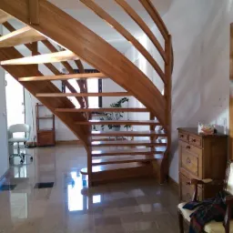 Jasne wnętrze z drewnianymi schodami spiralnymi, polerowaną podłogą i drewnianą stolarką drzwiową, oświetlone naturalnym światłem wpadającym przez okno.