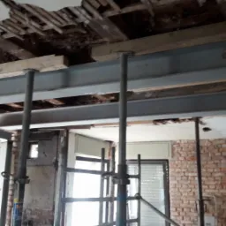 Pracownik w kasku mocuje stalową belkę pod zniszczonym stropem, widoczne rusztowania i odsłonięta ceglana ściana.