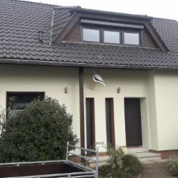 Jasna elewacja domu jednorodzinnego z brązowymi oknami i dachem z ciemnej dachówki, ozdobiona bocianem nad wejściem, widoczne krzewy i przyczepka.