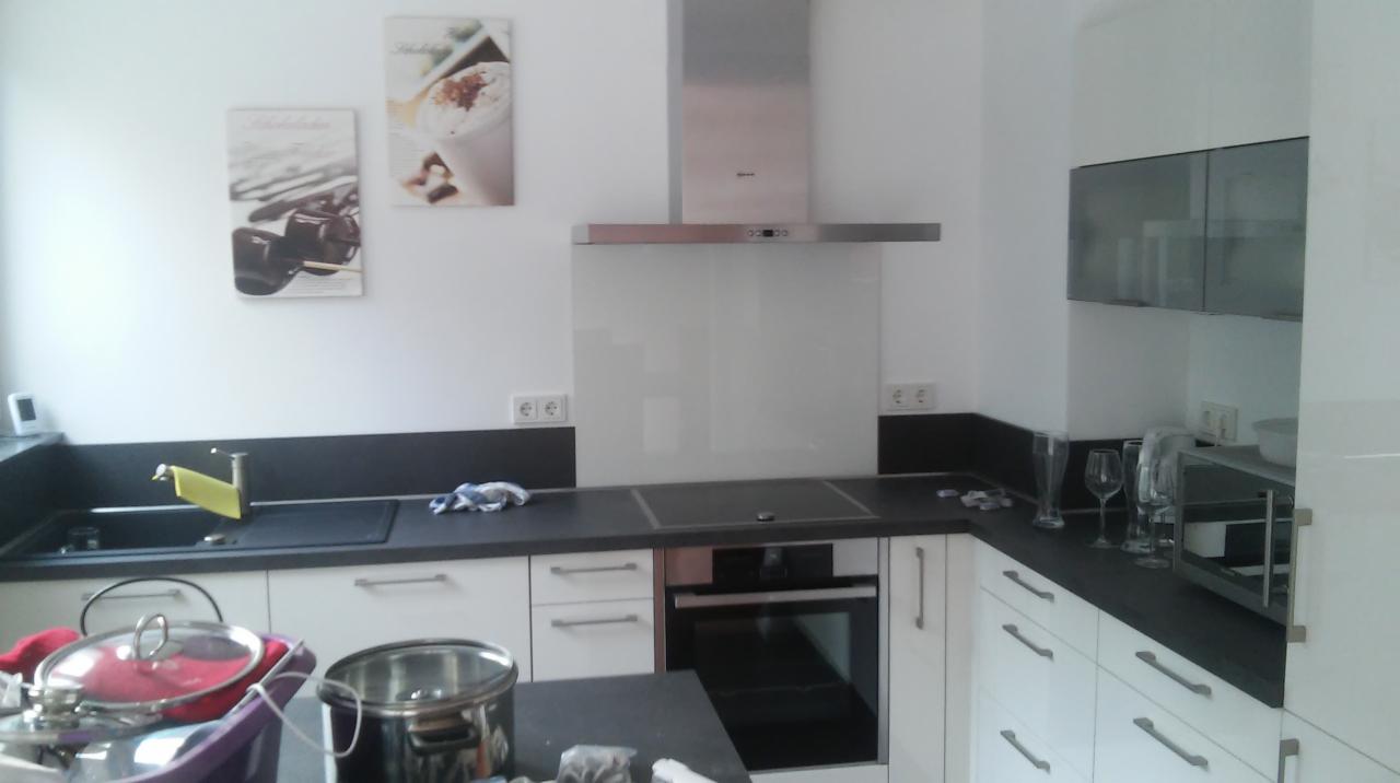 Nowoczesna kuchnia z białymi szafkami, czarnym blatem, szklanym panelem nad blatem i srebrnym okapem. Na ścianie wiszą dwa obrazy z motywami kawowymi.