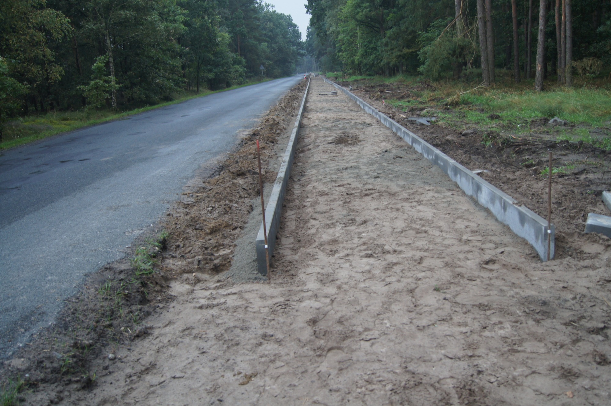 Przygotowanie podłoża pod chodnik z obrzeżami betonowymi wzdłuż asfaltowej drogi, widoczne wbite pręty stabilizujące.