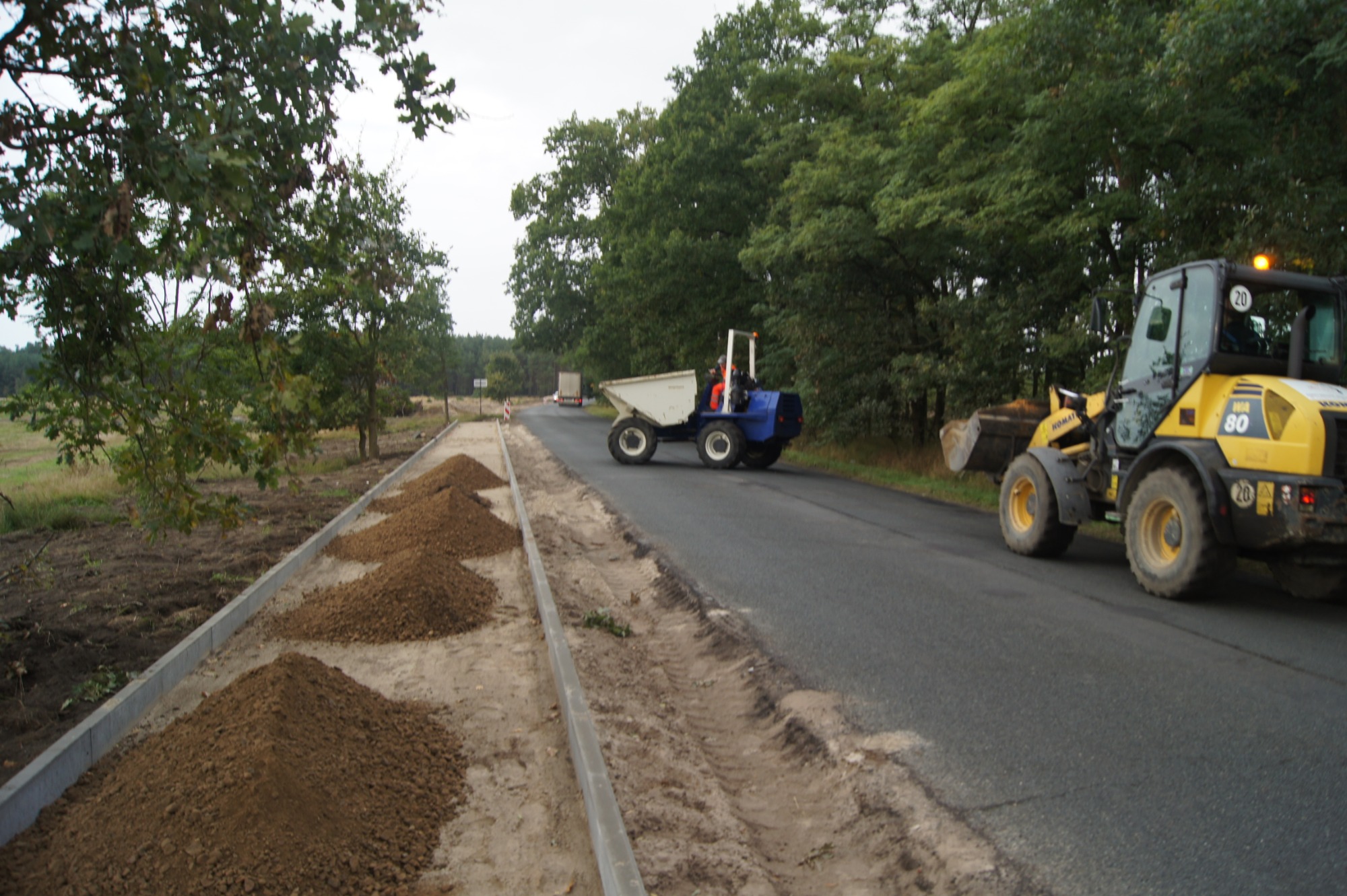 Żółta ładowarka i niebieska wywrotka na asfalcie obok pryzm ziemi i betonowych krawężników, w tle drzewa i przejeżdżający samochód.