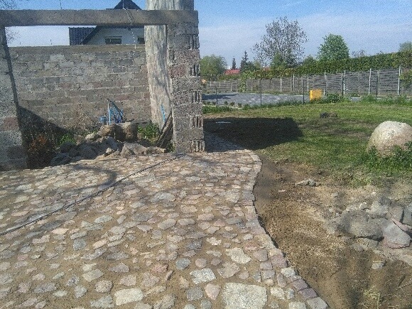 Fragment ścieżki z nieregularnej kostki granitowej, prowadzącej od niedokończonej konstrukcji budynku w stronę trawnika z dużym kamieniem. Widoczny cień budynku na trawniku.
