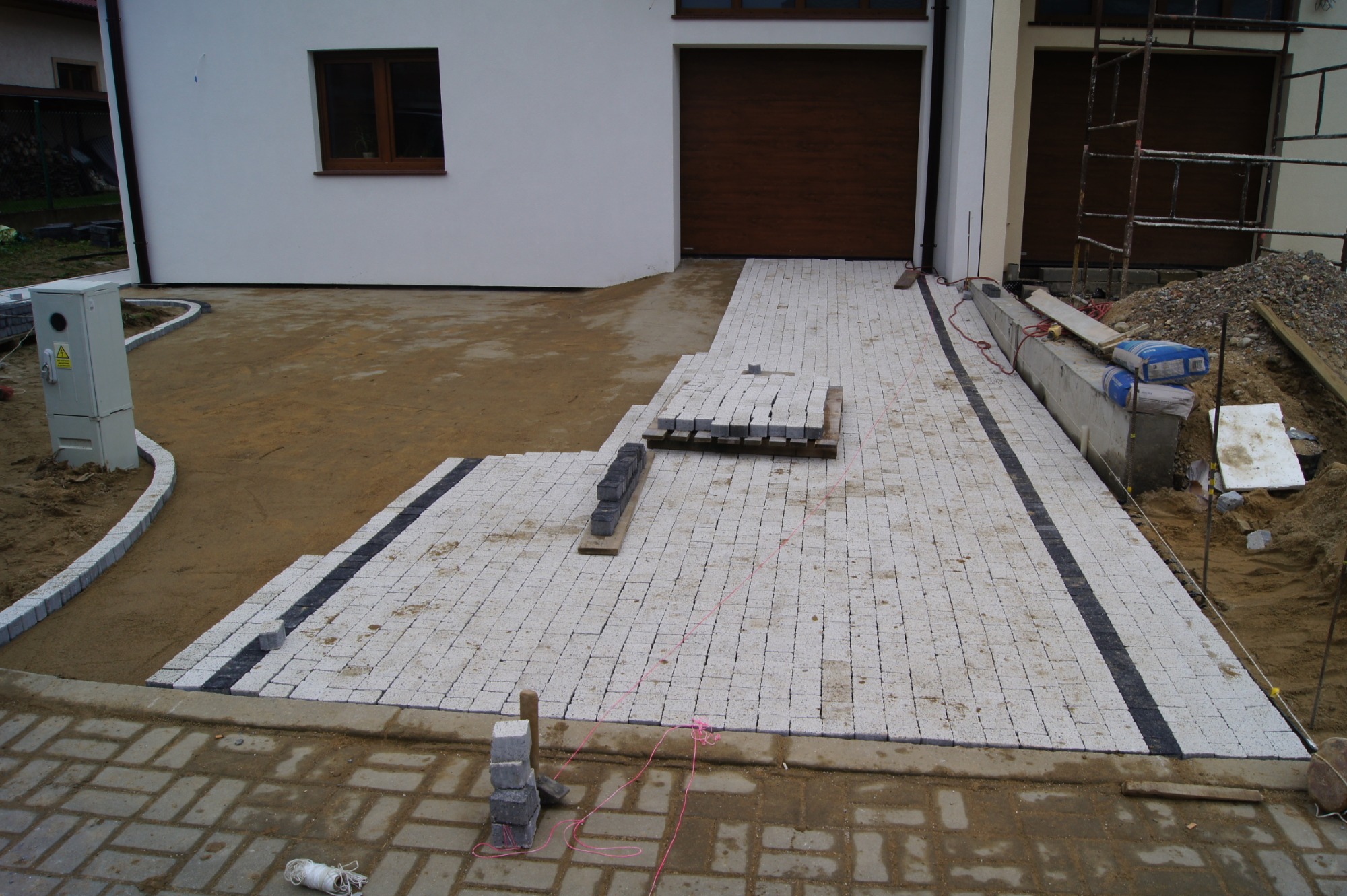Biała kostka ułożona częściowo na podjeździe, z czarnym pasem pośrodku, widoczne zapasy kostki na palecie, dom z brązową bramą garażową w tle, teren przygotowany pod dalsze prace.