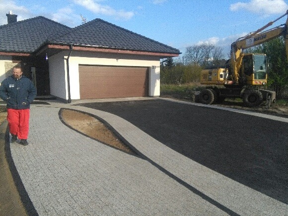 Ukończony podjazd do domu z kostki brukowej w odcieniach szarości i czerni, z wysepką ziemi w kształcie łzy, mężczyzna w czerwonych spodniach przechadza się po nowej nawierzchni, w tle dom...