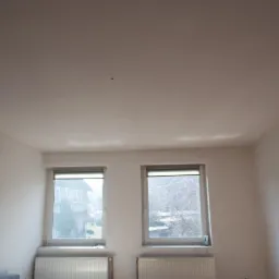 Pomalowane na biało wnętrze pokoju z dwoma oknami i grzejnikami pod nimi, widoczne ślady po malowaniu na suficie i ścianach.