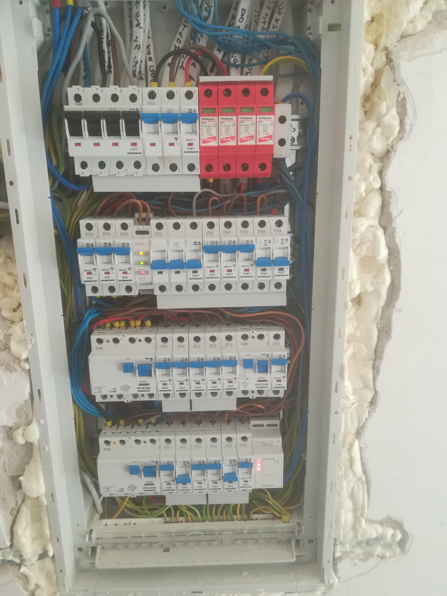 Rozdzielnica elektryczna z zainstalowanymi bezpiecznikami i ogranicznikami przepięć, widoczne okablowanie i pianka montażowa wokół obudowy, jeden z bezpieczników sygnalizuje stan włączenia diodą LED.
