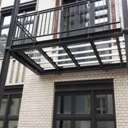 Metalowa konstrukcja balkonu z prostą, czarną barierką, zamontowana na elewacji z białej cegły z ciemnymi oknami.