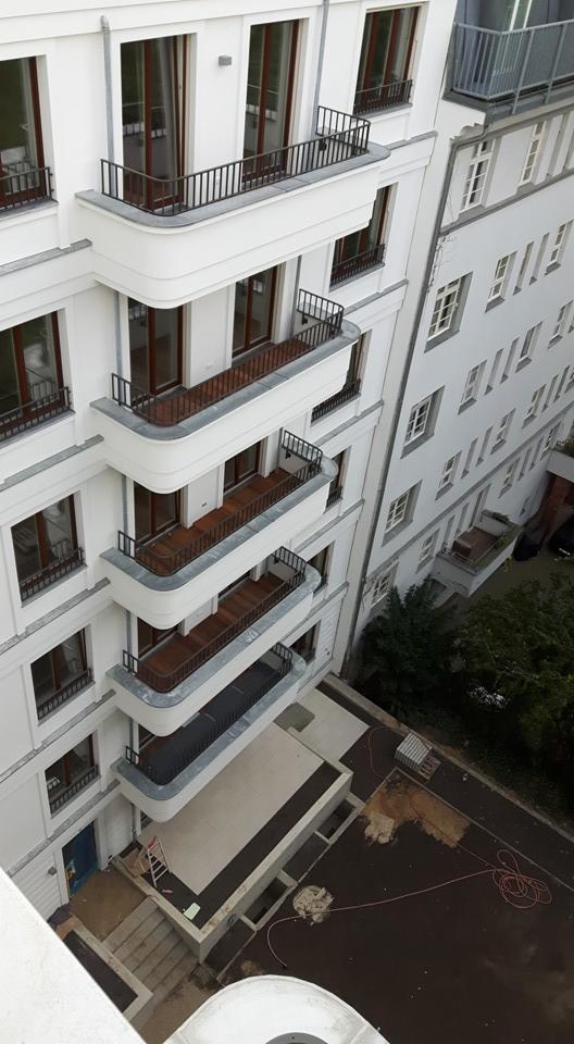 Elewacja budynku z balkonami zabezpieczonymi czarnymi, metalowymi barierkami, widok z góry na dziedziniec z rozłożonym wężem ogrodowym i drabiną opartą o ścianę.