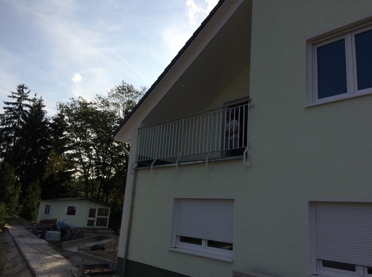 Balkon z nowoczesną, metalową konstrukcją zabezpieczającą, widok z zewnątrz na jasny budynek z roletami w oknach i fragmentem ogrodu w tle.