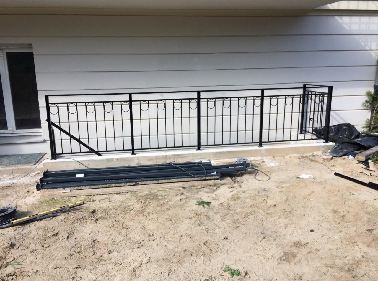 Czarna balustrada metalowa z dekoracyjnymi elementami owalnymi, zamontowana przy białej ścianie budynku. Na ziemi leżą rury i narzędzia, co sugeruje trwający montaż.