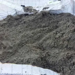 Worek typu Big Bag wypełniony piaskiem szarym, widoczne szwy worka, żółta etykieta i fragment czarnej folii wystające spod piasku.