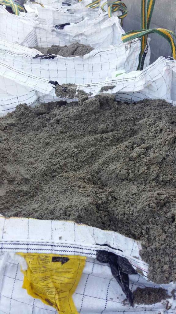 Worek typu Big Bag wypełniony piaskiem szarym, widoczne szwy worka, żółta etykieta i fragment czarnej folii wystające spod piasku.