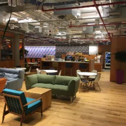 Przestronne wnętrze biurowe z drewnianą podłogą, sofami w odcieniach zieleni i błękitu, stolikami kawowymi, oraz barem w tle z ekspresami do kawy i automatem z napojami.